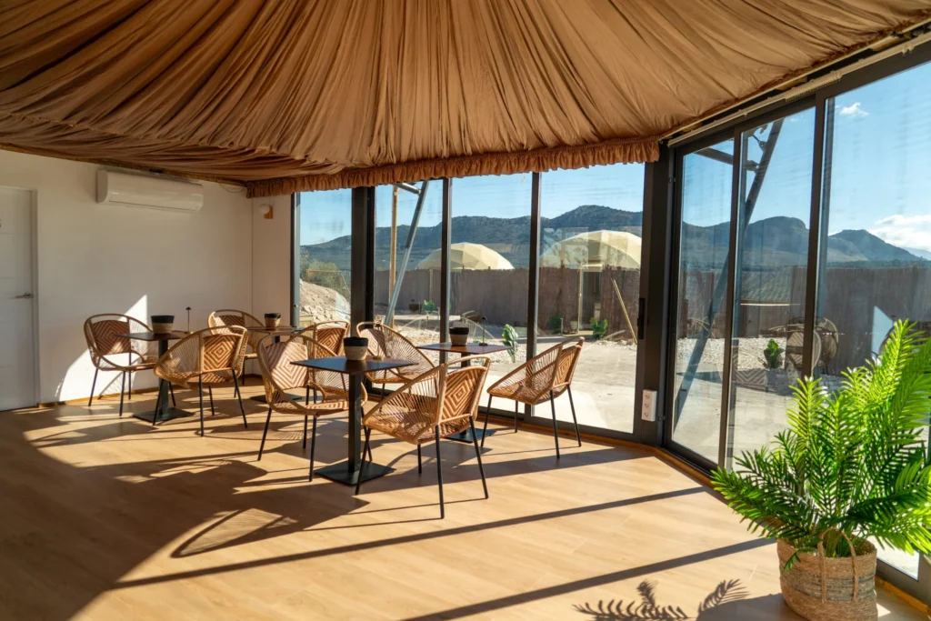 Salón comedor acristalado con techo de tela drapeada, mesas y sillas de mimbre, y grandes ventanales con vistas a la montaña