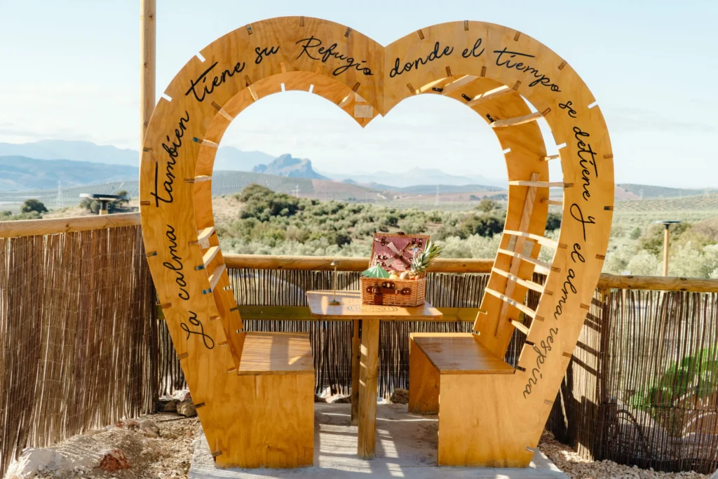 Estructura de madera en forma de corazón con bancos y cesta de picnic, situada en un mirador con vistas a la Peña de los Enamorados y olivares