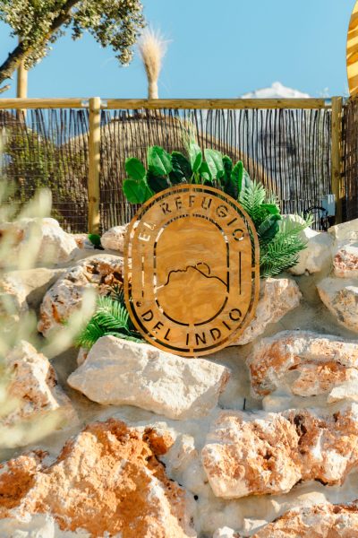 Cartel de madera con el logo de El Refugio del Indio sobre muro de piedra en la entrada