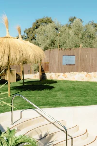 Zona de piscina con entrada escalonada tipo playa, césped artificial y sombrillas de paja estilo tropical en el exterior y arena de Tulum