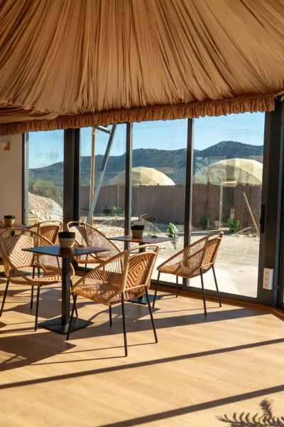 Salón comedor acristalado con techo de tela drapeada, mesas y sillas de mimbre, y grandes ventanales con vistas a la montaña